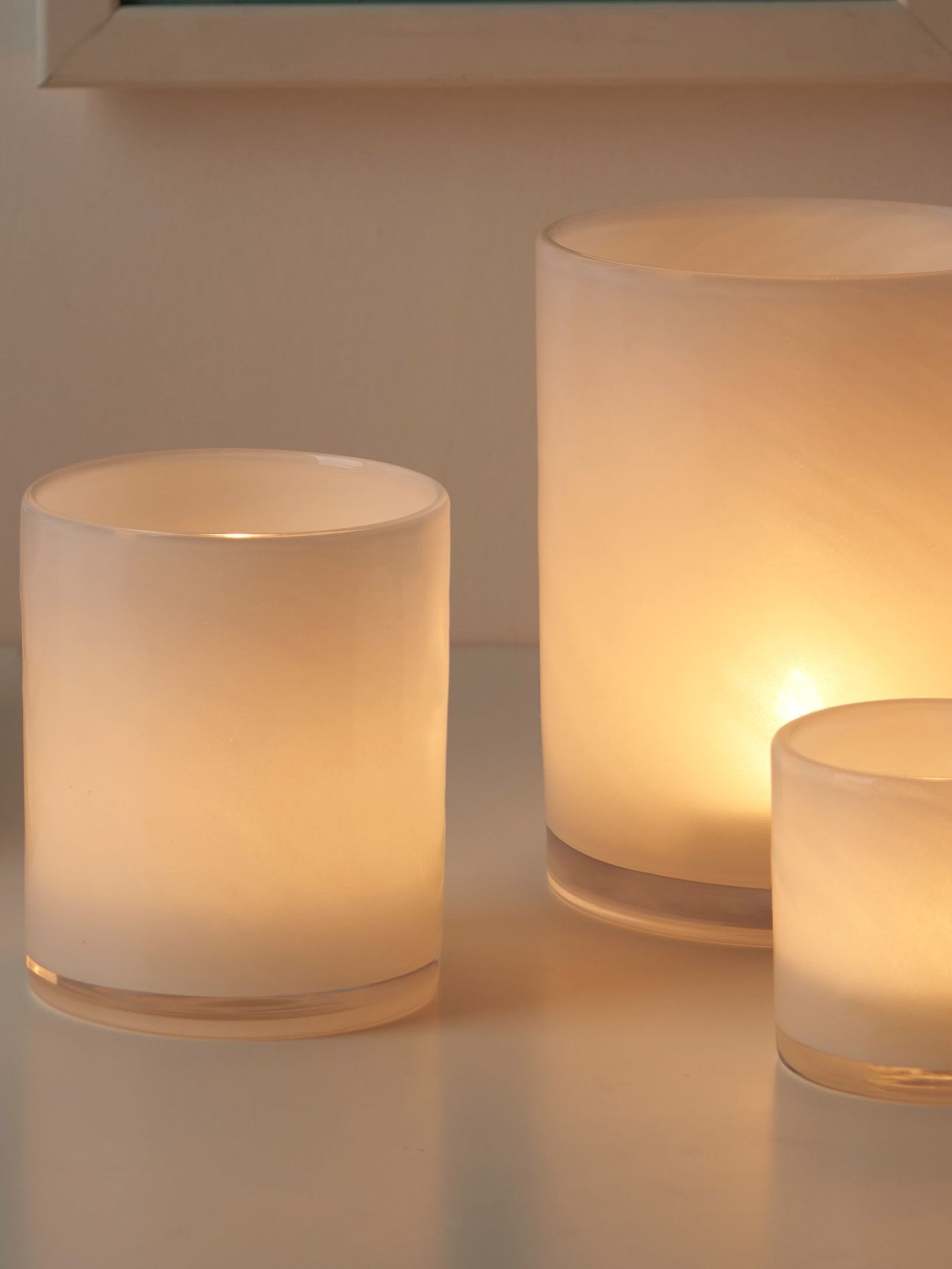 Närbild på tre stycken VINDSTILLA vita ljuslyktor med tända ljus i olika storlekar.