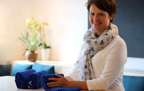 Person in hellem Raum arrangiert blaue gefaltete Stoffe auf einem Tisch; im Hintergrund stehen Kissen und eine Pflanze in warmem Licht.