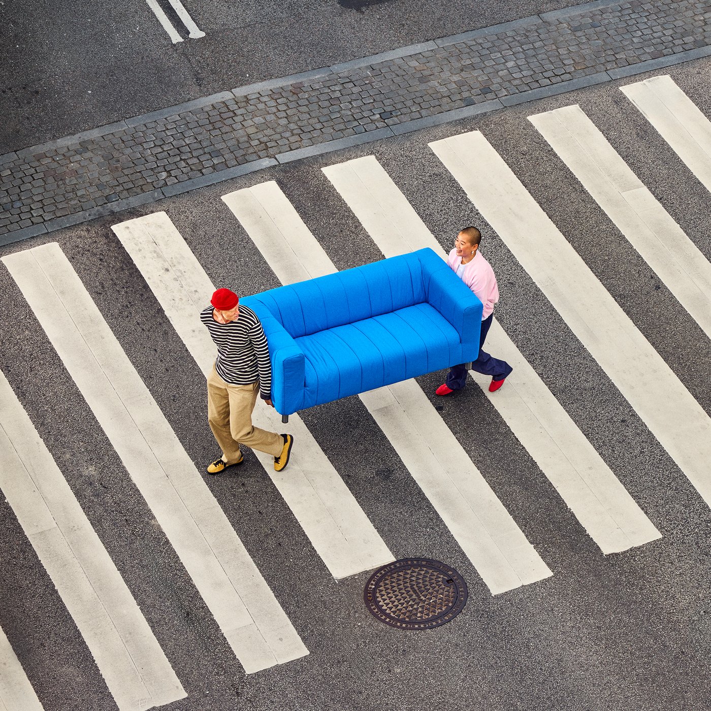 Two people carrying a blue KLIPPAN sofa across a crosswalk.