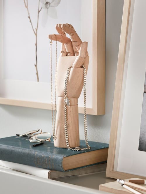 Between two frames with art stands a solid-beech HANDSKALAD decoration hand, a necklace draped over its thumb.