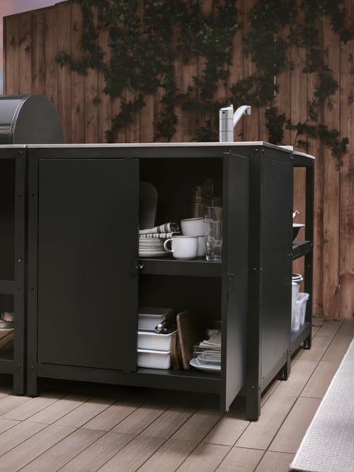 On a patio, an open door on a GRILLSKÄR kitchen island shows plates, cups, and serving dishes on the shelves inside.