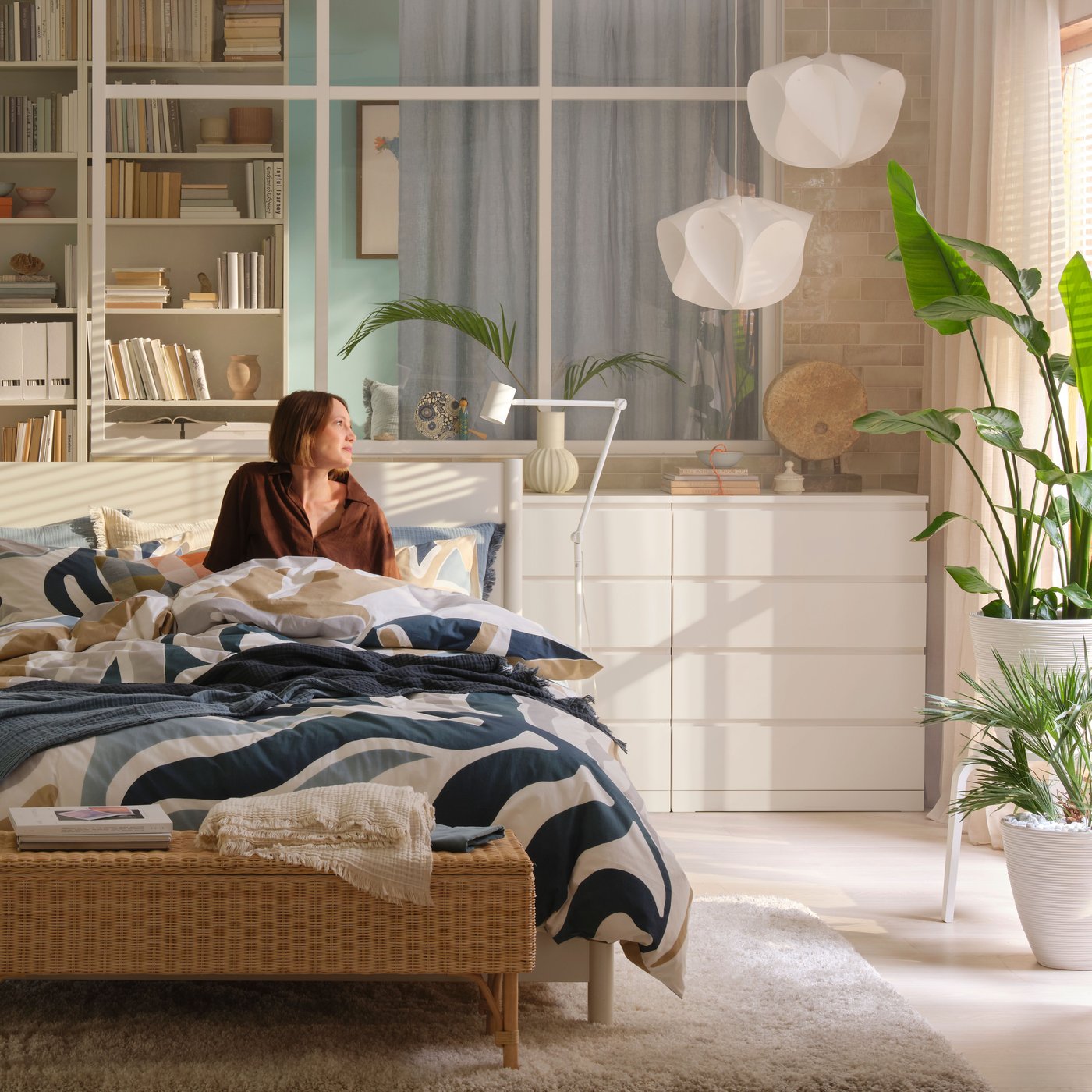 A woman sitting on a bed with blue and white bedsheet in a bedroom with white chest of drawers and white ceiling lamps