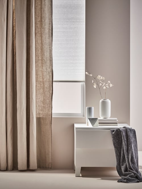 Ein Schlafzimmer mit Blick auf eine PLATSA Kommode mit 2 Schubladen vor einem Fenster. Aus der oberen der Schubladen hängt ein INGRUN Plaid heraus, auf ihr steht eine STILREN Vase. Am Fenster ist eine HOPPVALS Wabenjalousie zu sehen.
