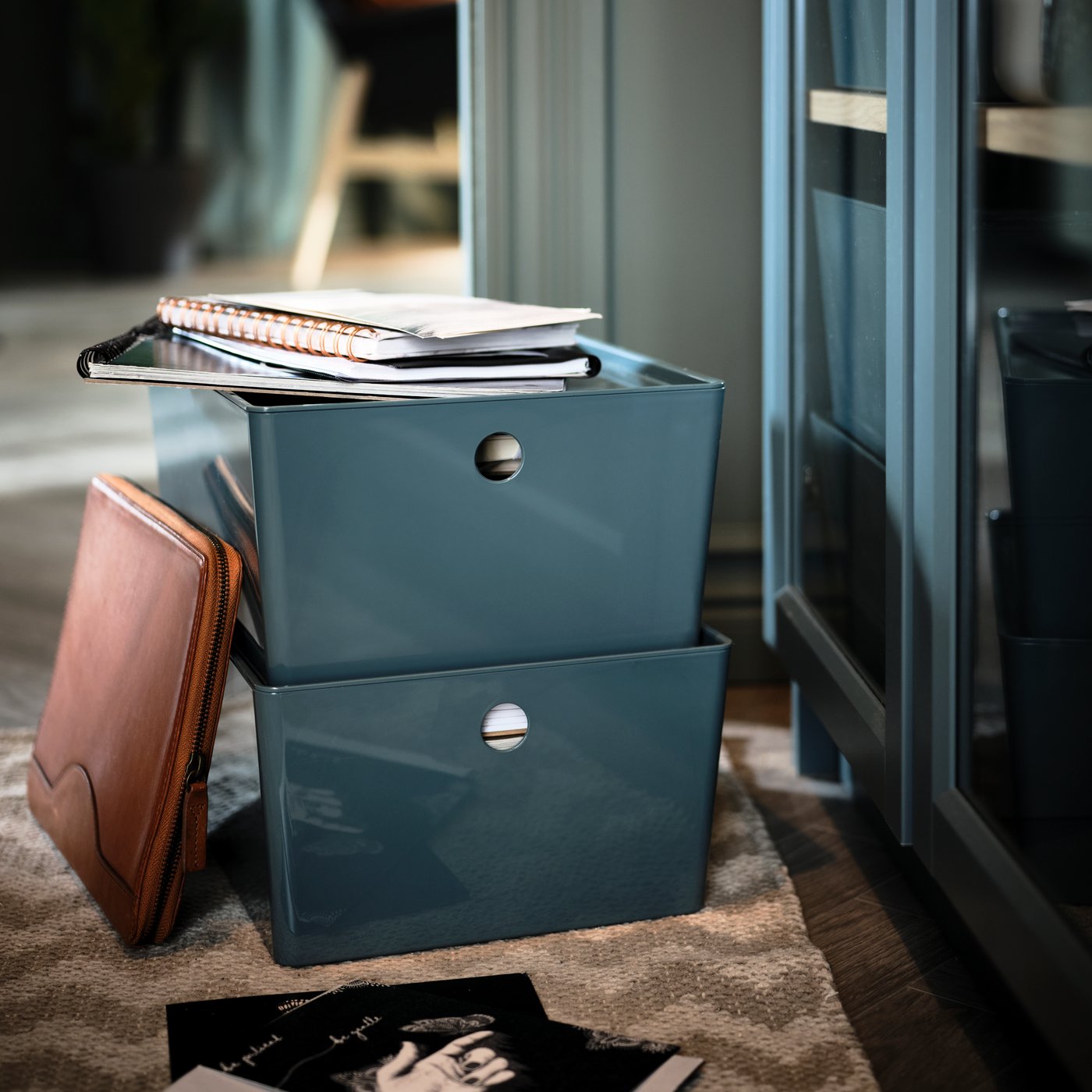 Two stacked, turquoise KUGGIS storage boxes holding notebooks and paper stand on a rug beneath a BILLY glass-door bookcase.