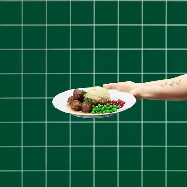 Käsi pitelee valkoista lautasta, jossa on HUVUDROLL-lihapullia, perunamuusia, herneitä ja puolukkahilloa. Taustalla on vihreä ruudullinen seinä.