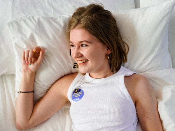 Izzy liegt auf einem Bett in einem weissen T-Shirt. Sie schaut nach rechts und lächelt, während sie ihre rechte Hand an ihr Gesicht hält.
