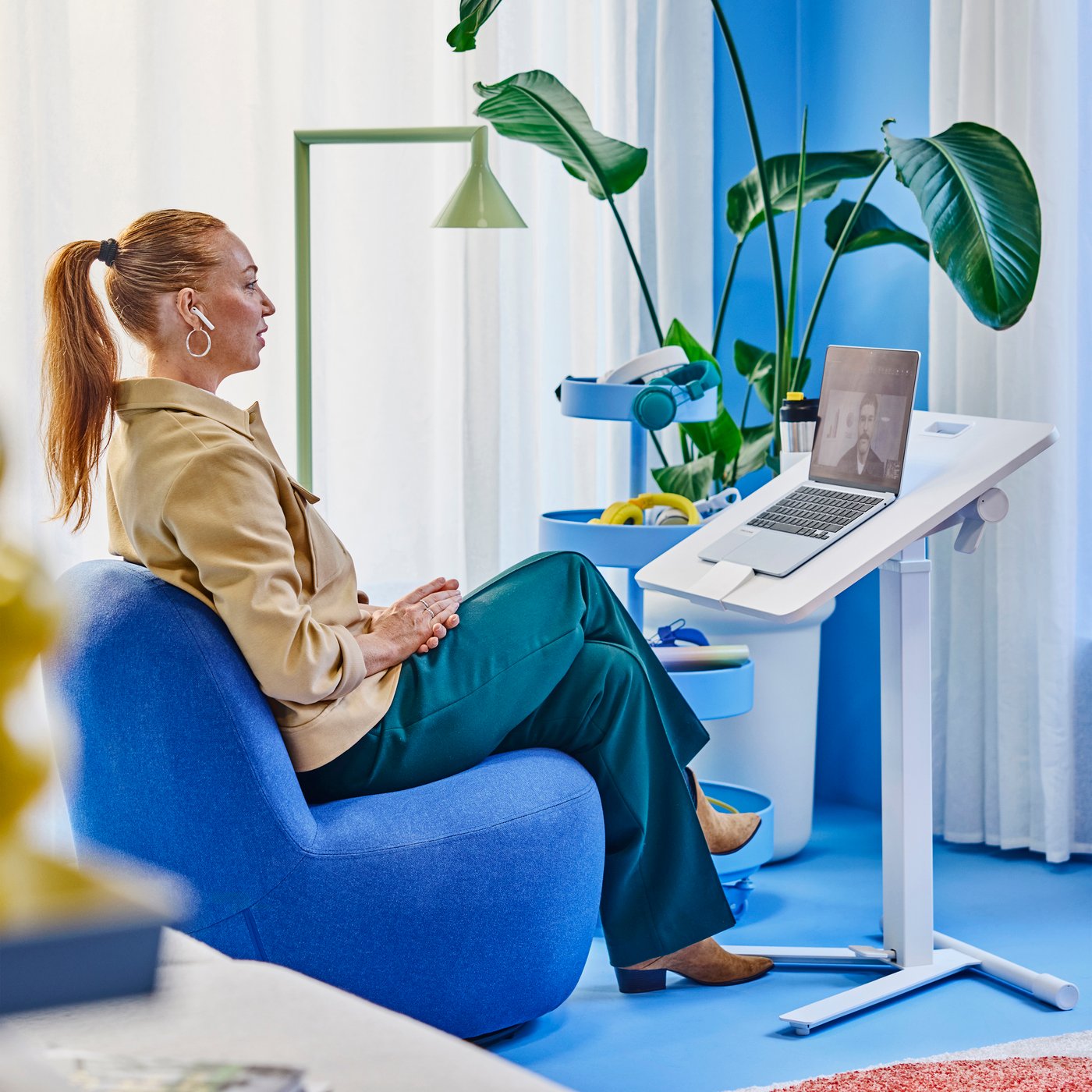 Person sittande i blå stol använder bärbar dator på justerbart skrivbord, med växt, lampa och tillbehör i ljust, modernt rum.