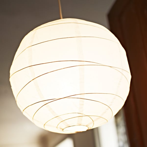 A white REGOLIT pendant lamp, which has a lit light bulb inside it, hangs from a ceiling in a living room.