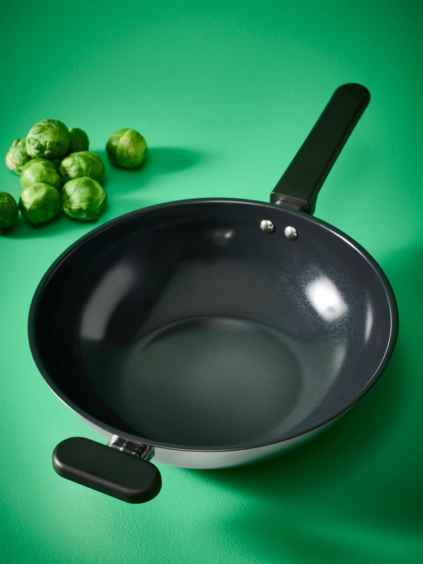 A MIDDAGSMAT wok in stainless steel placed on a green surface next to some brussels sprouts.