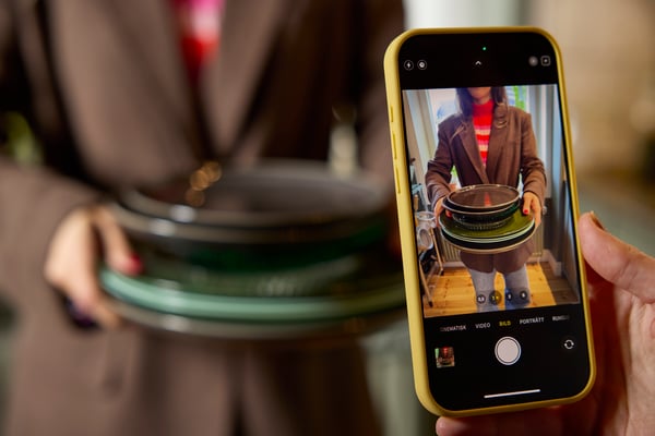 Primer pla d'un mòbil fent una foto a una persona que sosté diversos plats apilats. Imatge que mostra com fotografiar objectes de la llar per vendre'ls o compartir-los en línia.