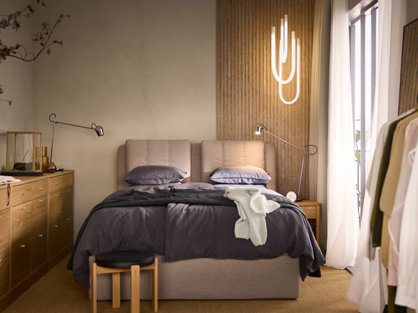 En LILLFJÄLLET kontinentalseng med opbevaring med mørkegråt sengetøj ved en beige væg med en VARMBLIXT LED-lampe hængende over.