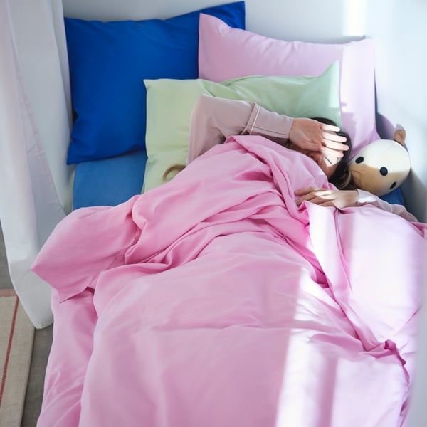 A person lying in a bed with the light pink PILTANDVINGE duvet cover and pillows in light green and blue, sunlight coming in.
