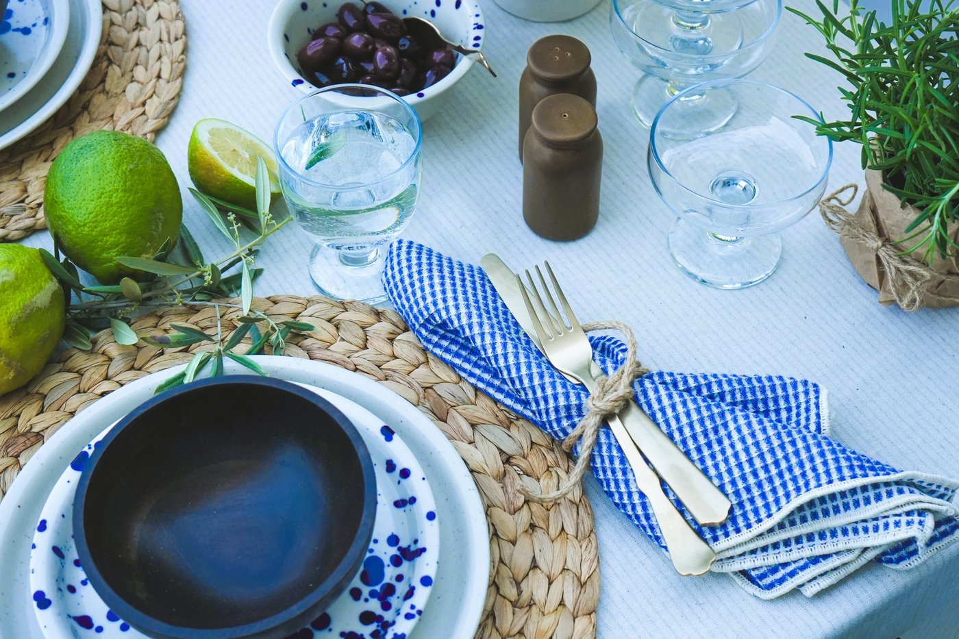 Table dressée avec des assiettes IKEA Silvereda, des couverts NATTKLOT, une serviette bleue et blanche, des citrons verts et des olives. Idéal pour les repas estivaux à la méditerranéenne.