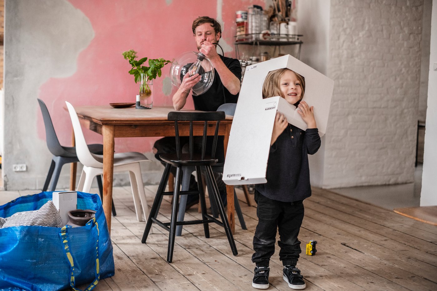 A father who is assembling a lamp at the dining table while his son plays with the cardboard box.