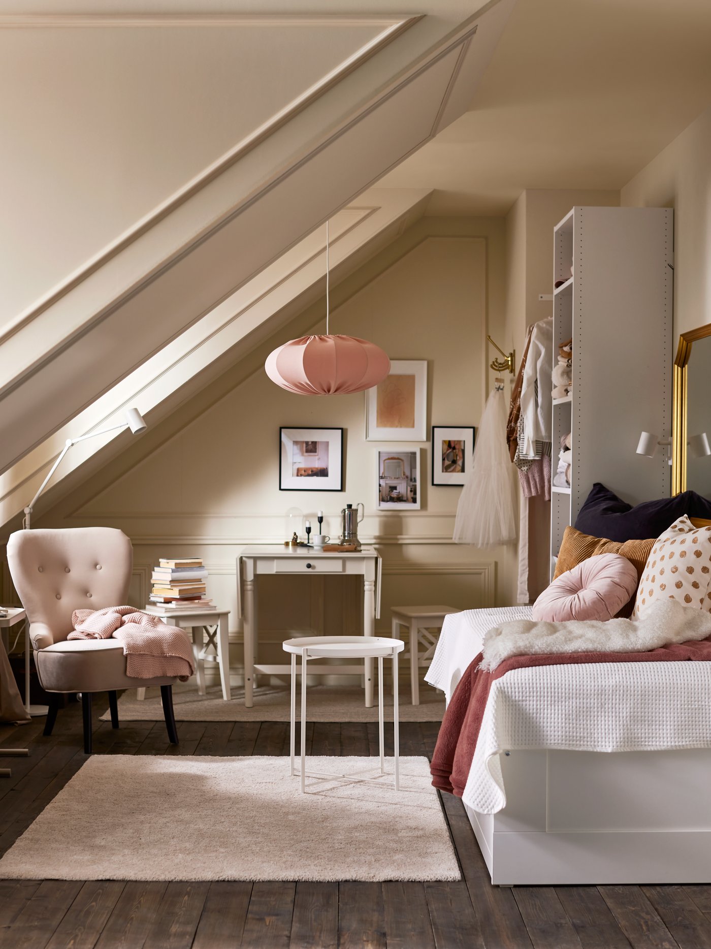 Dormitorio con una mesa con bandeja redonda, una estructura de diván con dos cajones, una lámpara de techo rosa, un escritorio pequeño y un sillón.