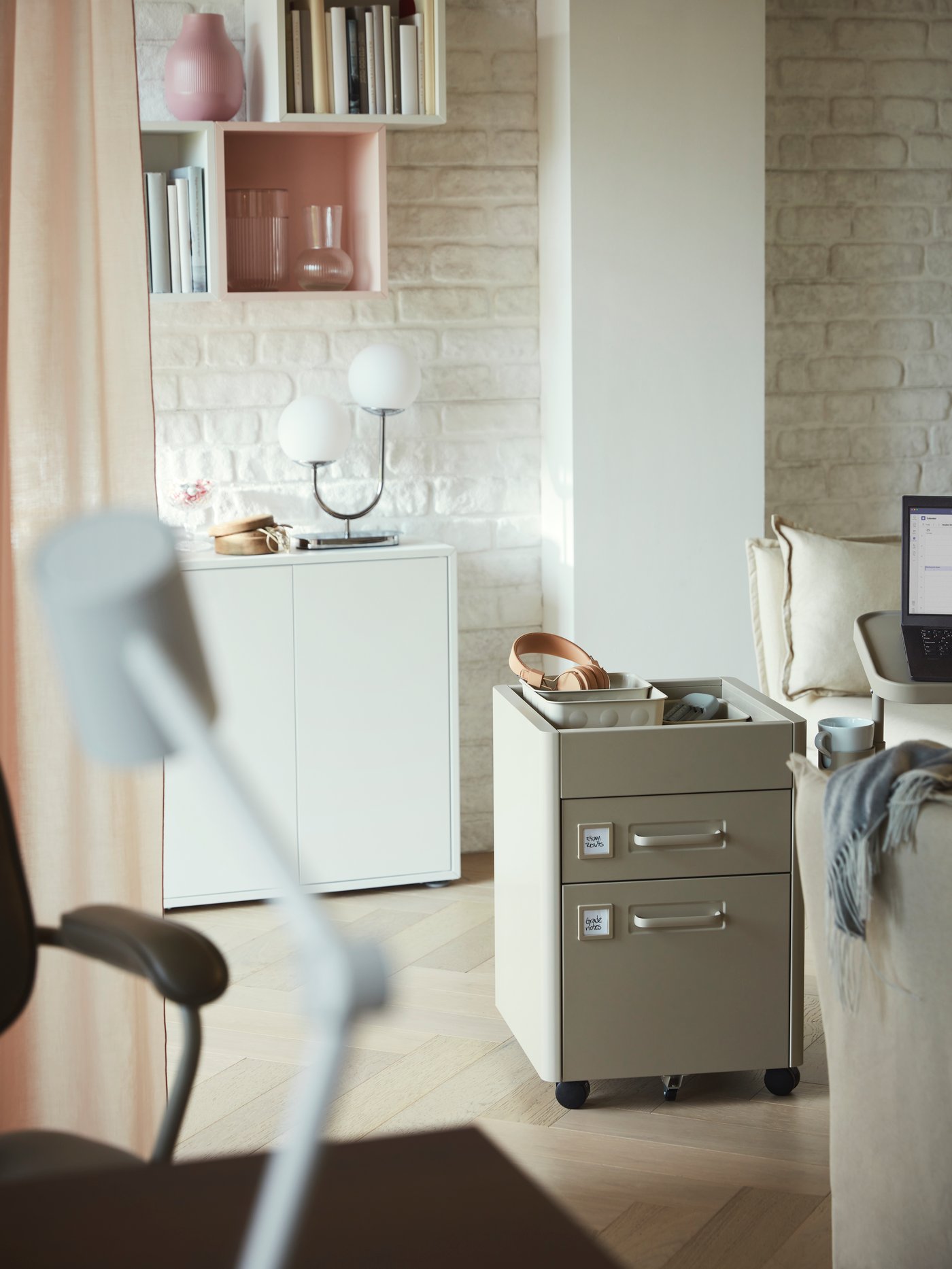A beige IDÅSEN drawer unit on castors near a sofa with a pair of headphones in a box on top in a white living room.