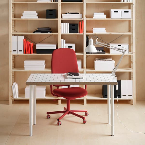A white LAGKAPTEN tabletop on OLOV adjustable legs, in front of a red office chair and IVAR shelves in pine.