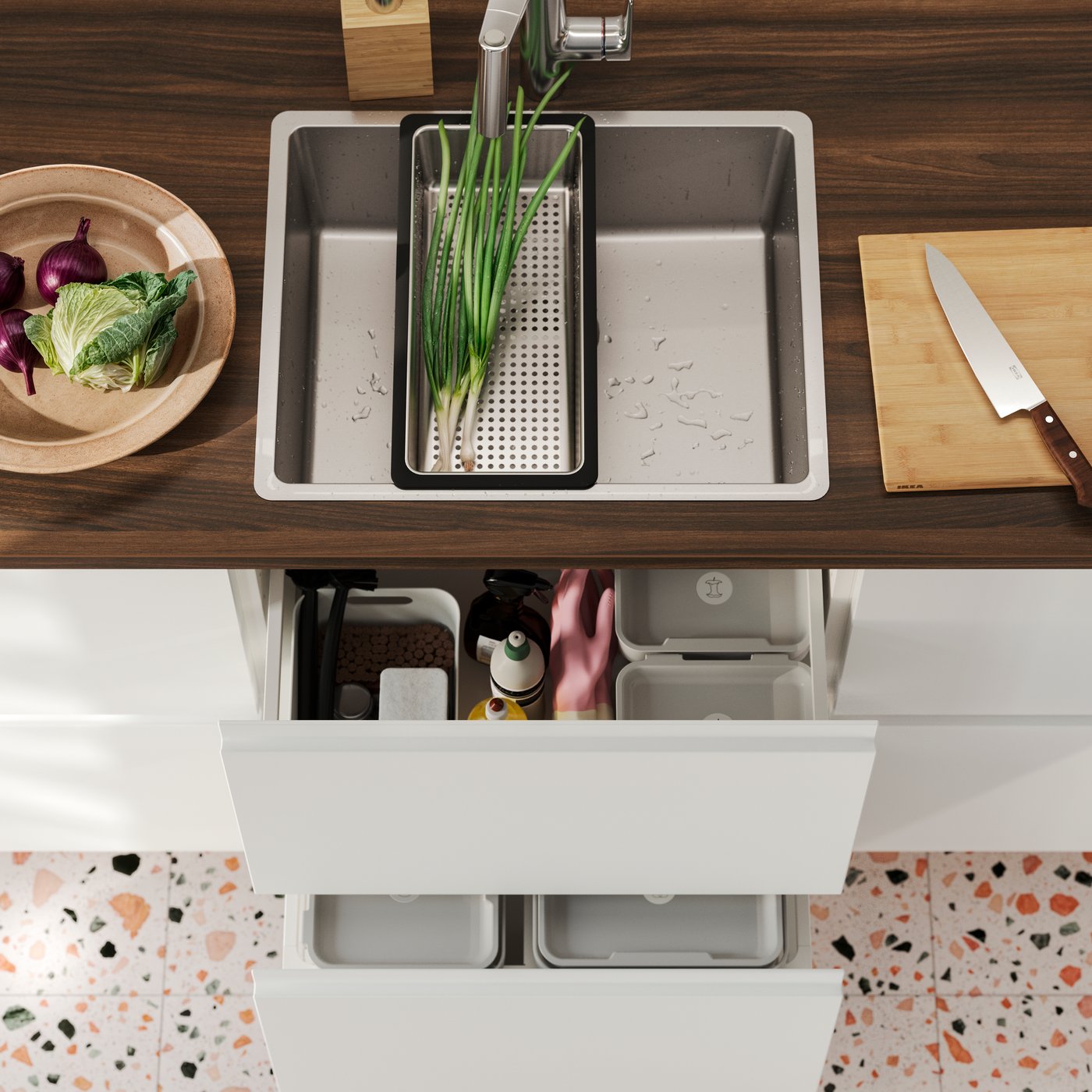 A VRESJÖN inset sink in stainless steel has a NORRSJÖN colander with some spring onions and next to it is a chopping board.