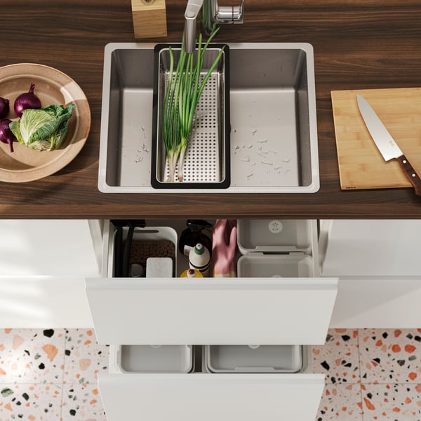 A VRESJÖN inset sink in stainless steel has a NORRSJÖN colander with some spring onions and next to it is a chopping board.