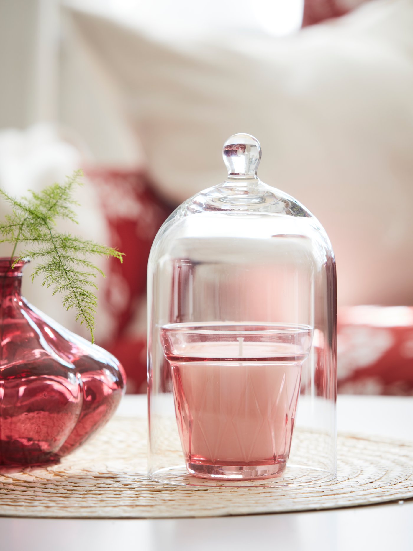 A glass dome covers a scented candle in a dark pink glass. Next to it is a small, dark red glass vase with a green twig.