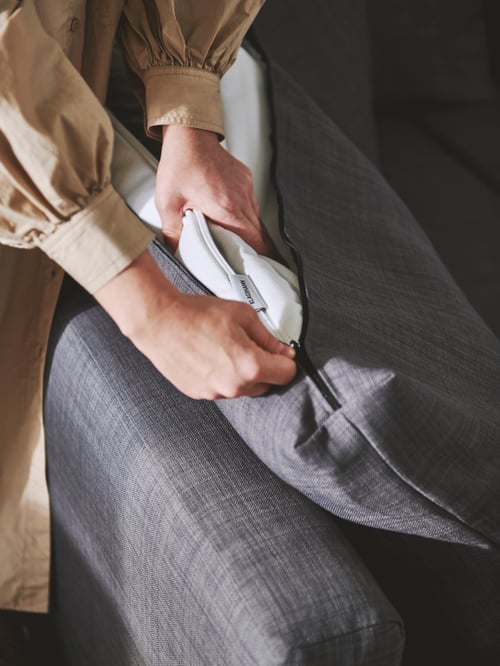 Close-up of a person's right hand holding the zipper of a KLAGSHAMN back cushion, while the left hand is holding the insert.