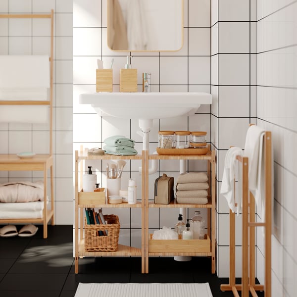 Two bamboo RÅGRUND wash-basin corner shelves filled with toiletries are under a sink in a bathroom. Towels hang on a rack.