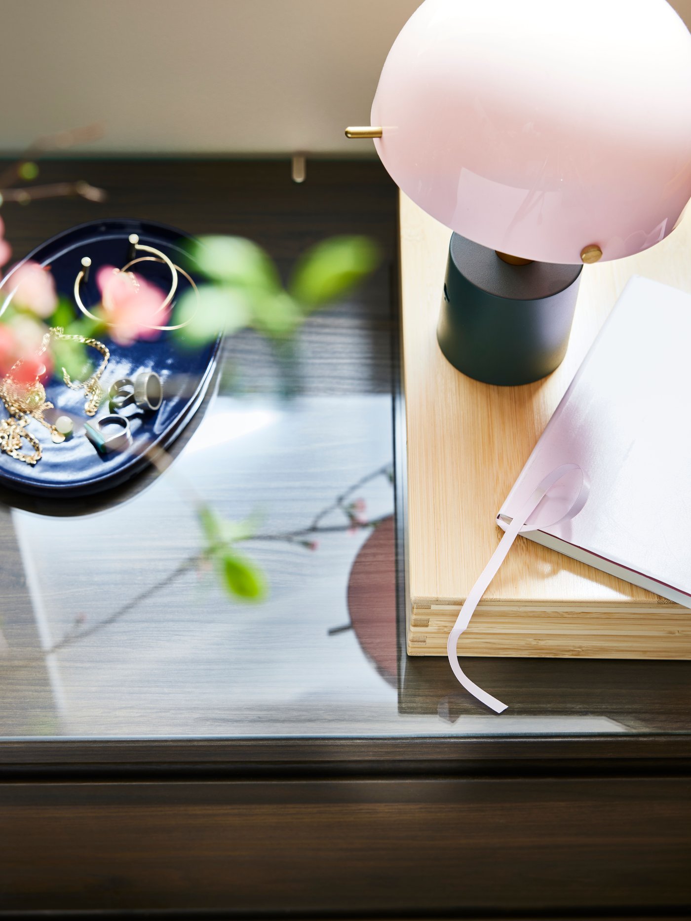 Een transparant STORKLINTA glazen blad bovenop een ladekast met daarop een roze/donkergrijsgroene NÖDMAST LED-draagbare lamp.