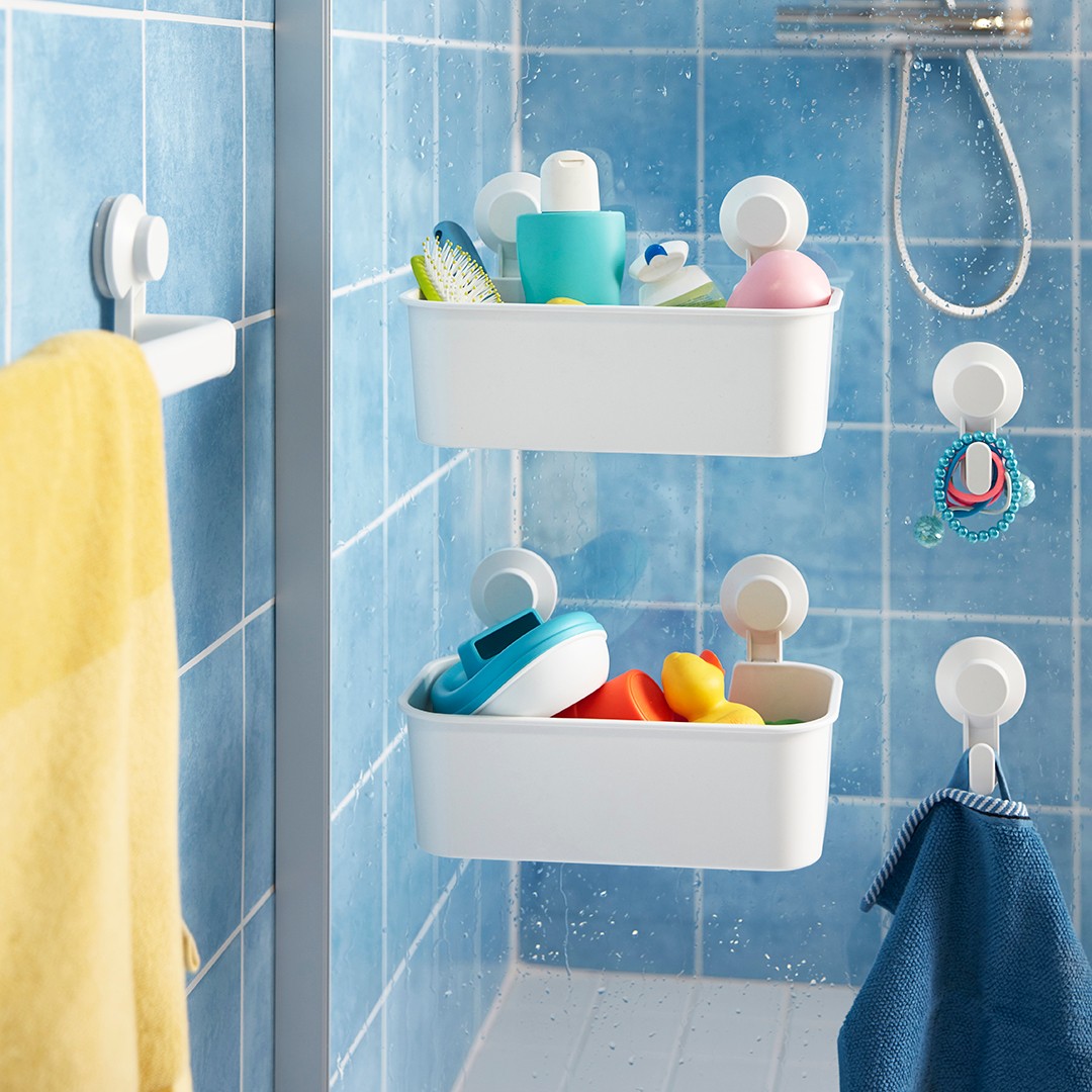 Vista de cerca de canastos plásticos TISKEN afirmados con ventosas a la mampara de baño. Se ven también perchas y un toallero TISKEN con una toalla amarilla colgada. Los muros del baño están revestidos en cerámicas celestes cuadradas con fragüe blanco.