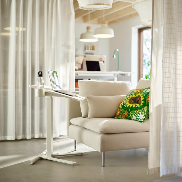 Single white sofa unit with deskstand holding a laptop. Communal workspace in background; divider curtains on either side.