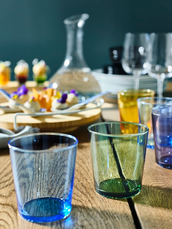 PAPPERSBJÖRK glasses, in blue, green and yellow, stand on a wooden table in front of a lazy susan with food.