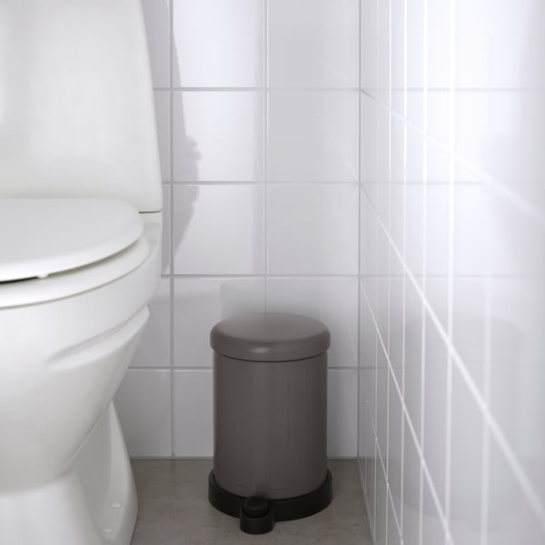 The image shows a section of an interior space, featuring a ribbed metal laundry basket with a curved top edge and a single gray handle on the side. The basket is placed on a teal-colored carpeted floor against a wall with vertical wooden paneling partially obscured by what appears to be the edge of a white door or divider. To the left, there is the partial view of another object with intricate patterns, possibly part of furniture or home decor. 