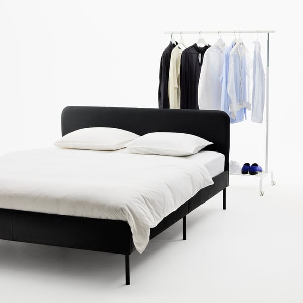 A black SLATTUM bed, neatly made with white bed linen. Behind the bed is a rack with colour-coordinated shirts and shoes.