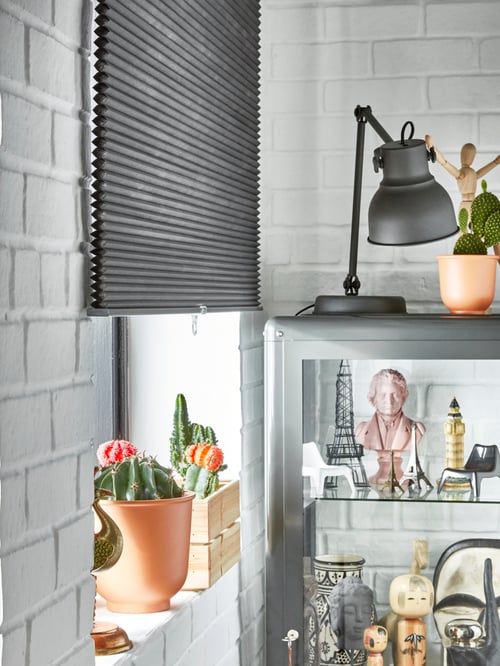 A grey HOPPVALS room darkening cellular blind covers half of a window by a glass-door cabinet filled with various trinkets.