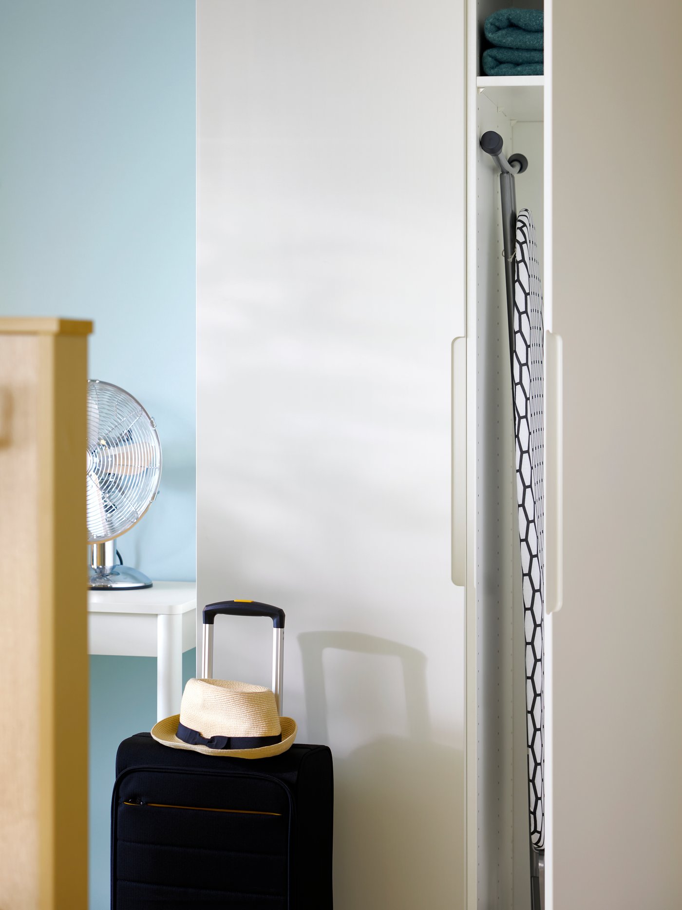 Armadio PAX bianco con ante TONSTAD lisce color bianco sporco, affiancato da una valigia pronta all’uso. Una delle ante è aperta e mostra l’asse da stiro all’interno, per tenere tutto in ordine e a portata di mano.