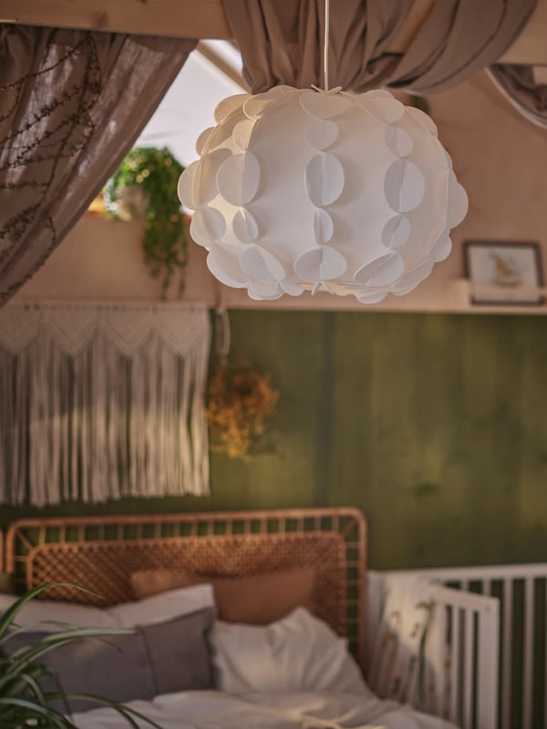 In a room with green and beige walls, a white PEKTOLIT pendant lampshade hangs above a VEVELSTAD bed and a GULLIVER cot.