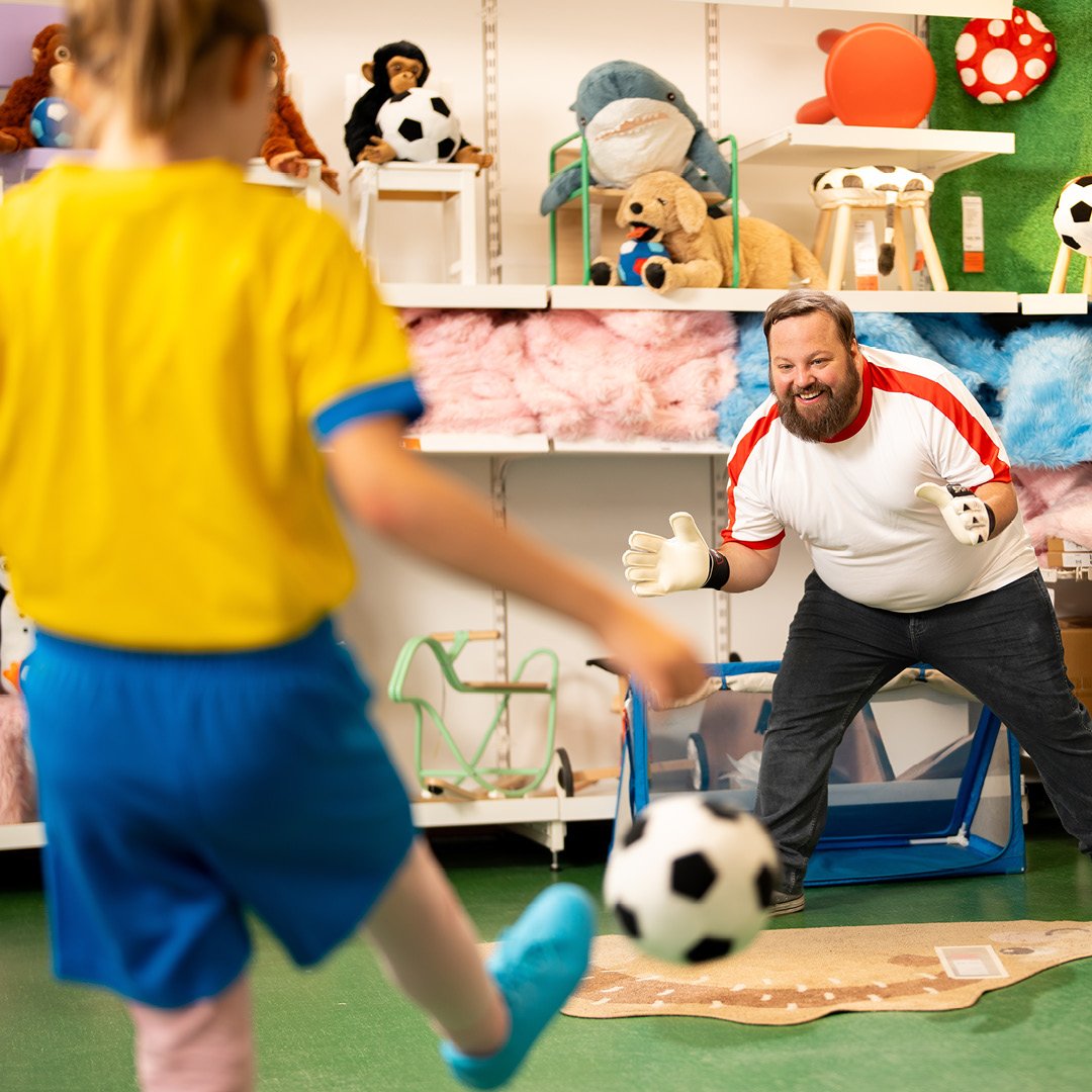 Eine Person mit einem Fußballtrikot versucht den Ball, den ein Kind in seine Richtung schießt, aufzufangen.