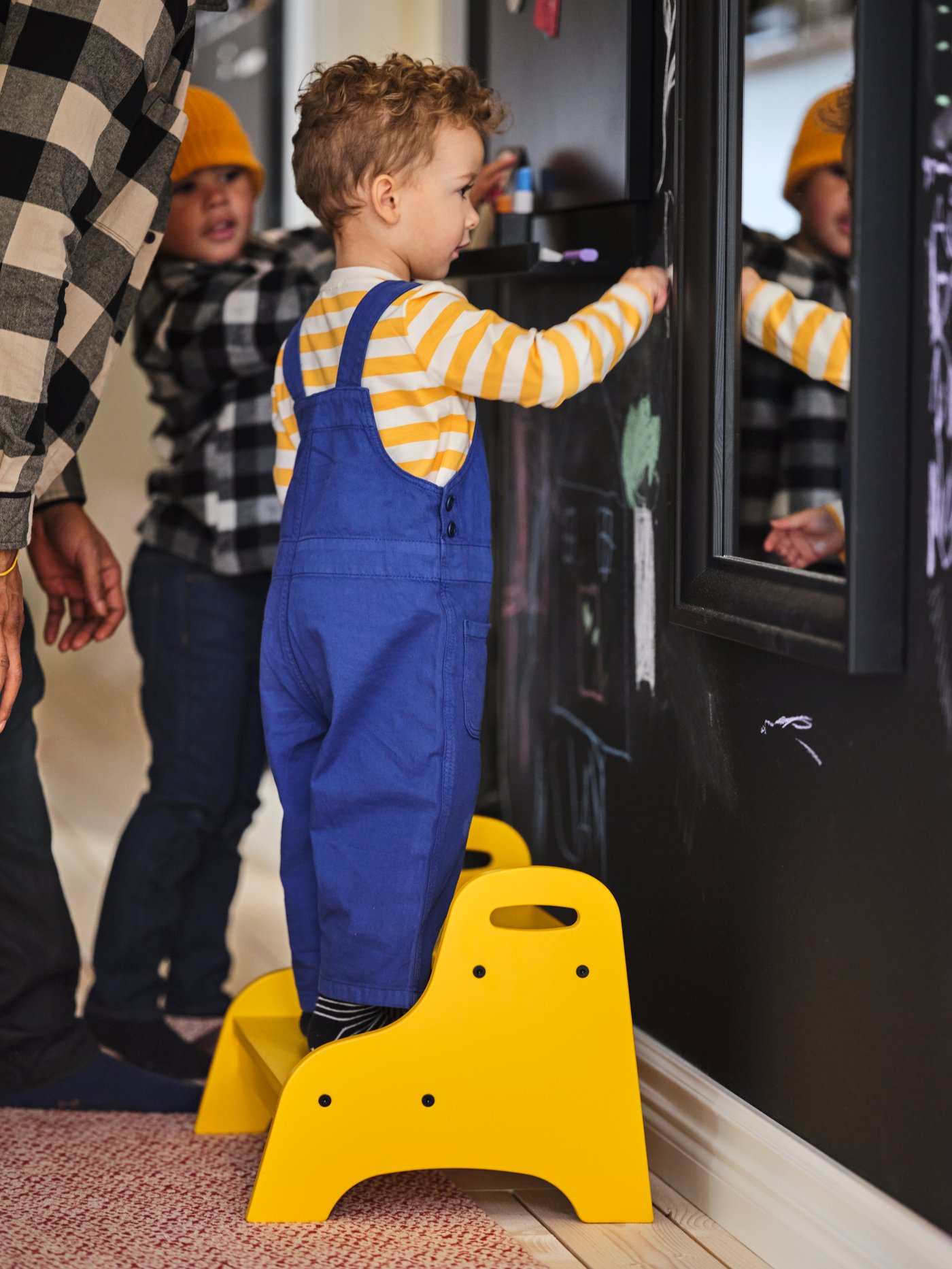 صبي يقف على سلم متدرج للأطفال ‪TROGEN‬ أصفر أمام مرآة ‪TOFTBYN‬ سوداء، وشقيقه ووالده على مقربة منه.
