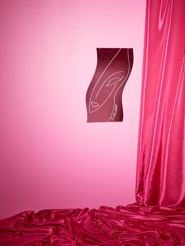 Ein KARISMATISK Spiegel, in den das Gesicht von Zandra Rhodes eingebrannt ist, hängt an einer Wand in Rosa neben einer rosafarbenen Gardine aus Satin. 