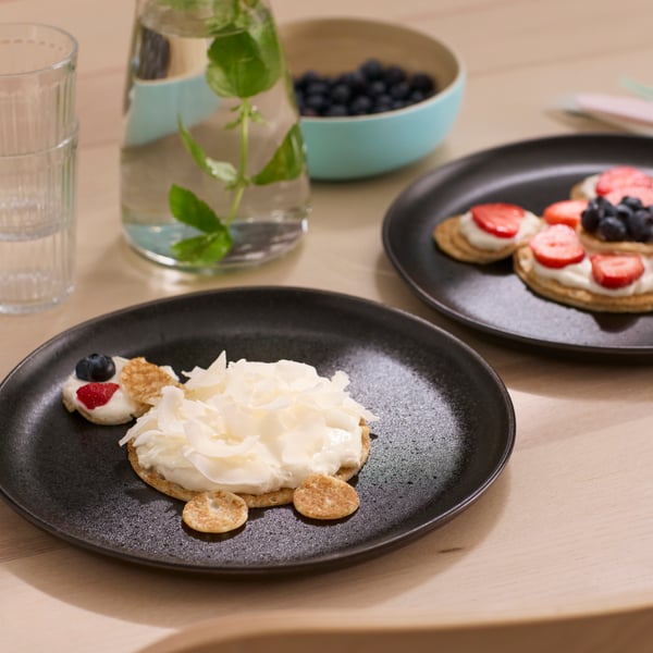A black plate with pancakes, whipped cream and fruit, cut in smaller pieces and laid out to look like a sheep.