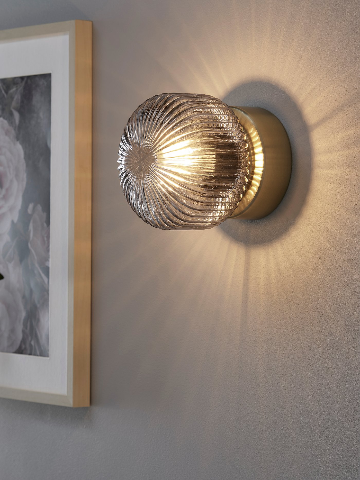 Een messing/grijs transparant glazen SOLKLINT wandlamp die een mooie schaduw geeft op de muur.