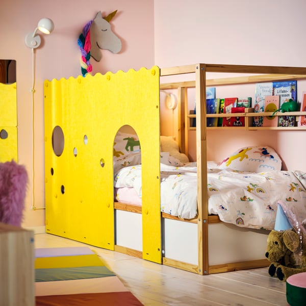 A KURA bed accessory in yellow with arched cut-outs on a pine bed, in a pink children’s room with colourful bedding.