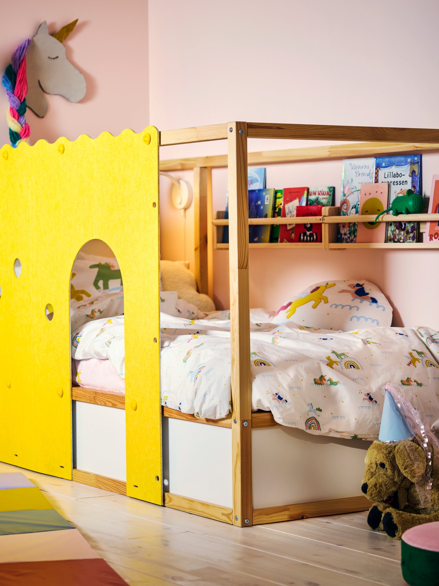 A KURA bed accessory in yellow with arched cut-outs on a pine bed, in a pink children’s room with colourful bedding.