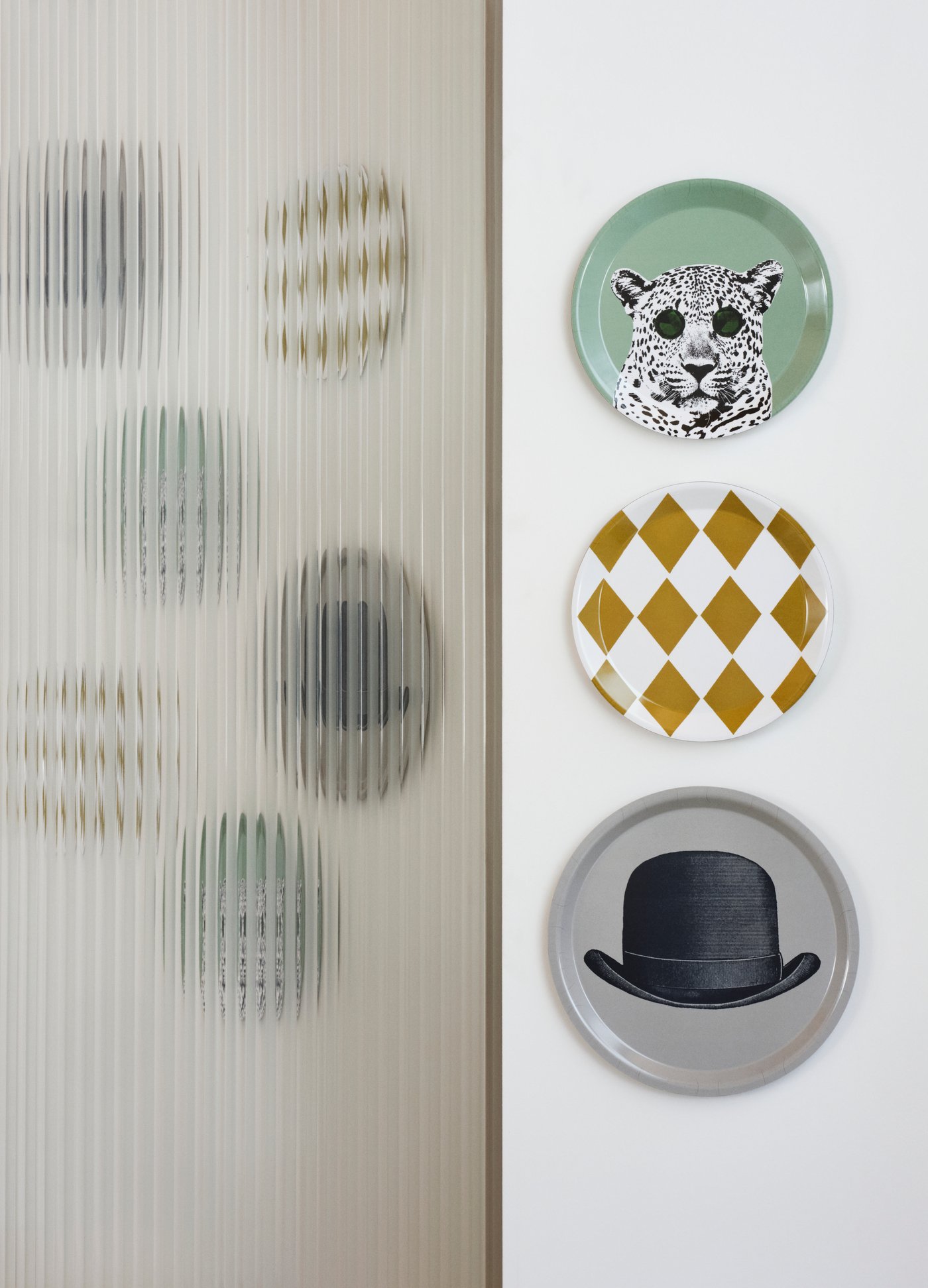 Tres platos decorativos de IKEA colgados de una pared. Uno tiene un guepardo, otro cuadros blancos y amarillos y el tercero un sombrero negro sobre un fondo gris.