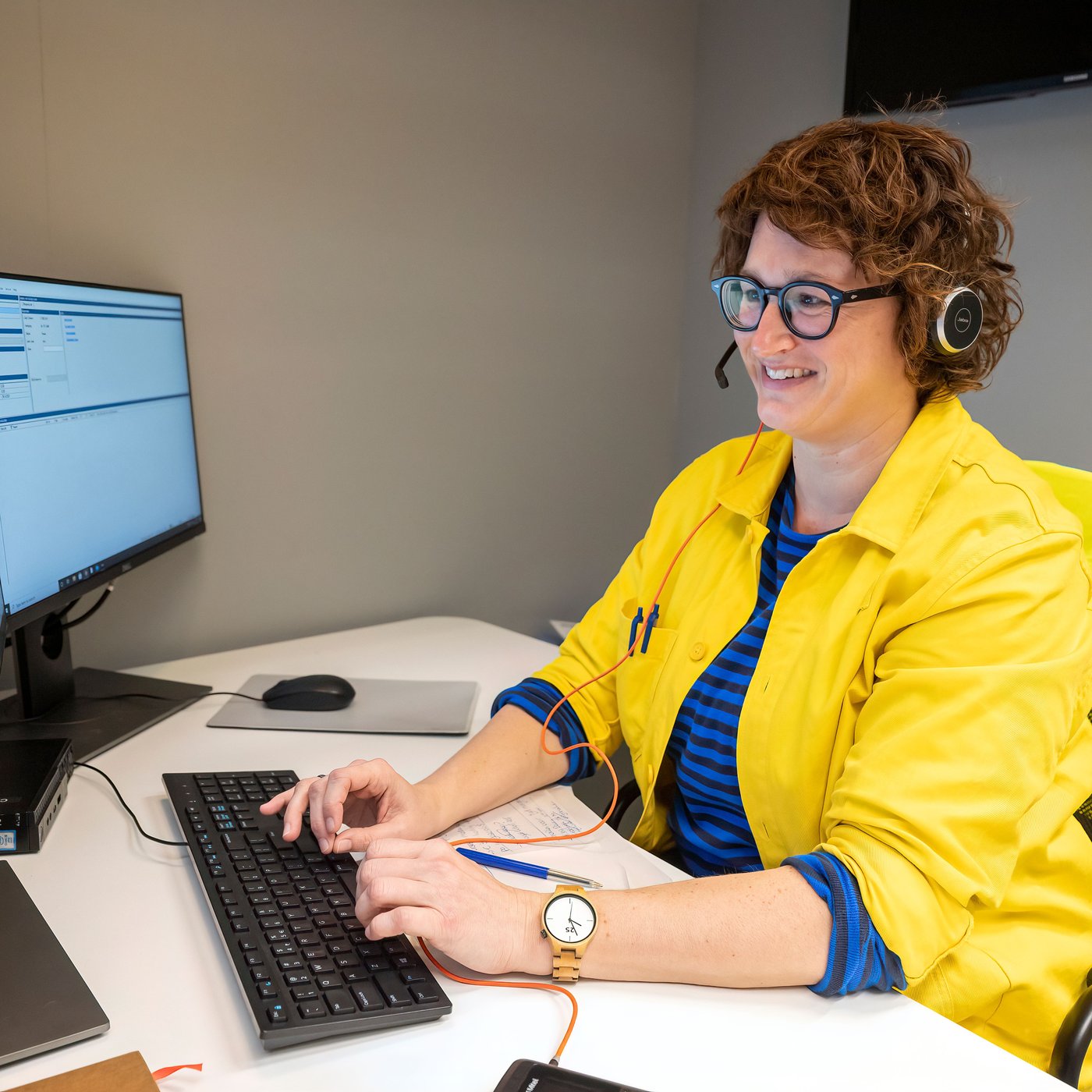 Persoon met headset werkt aan een bureau, typt op toetsenbord naast computerscherm met software-interface.