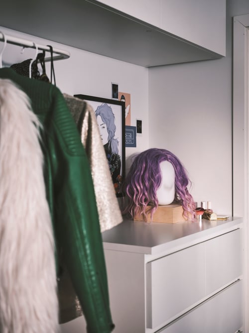 Bedroom area featuring a head-shaped table lamp with a purple wig set on a white chest. Next to the chest are clothes.