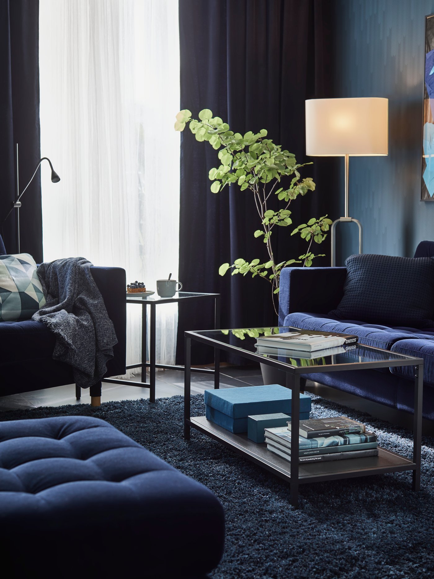 Un sofá azul, un reposapiés y un sillón con una mesa de centro VITTSJÖ de vidrio entre ellos y otra junto al sillón.