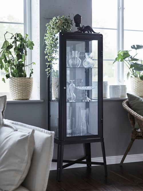A FABRIKÖR glass display cabinet between two windows filled with lots of glassware. Plants are on top and to the side.