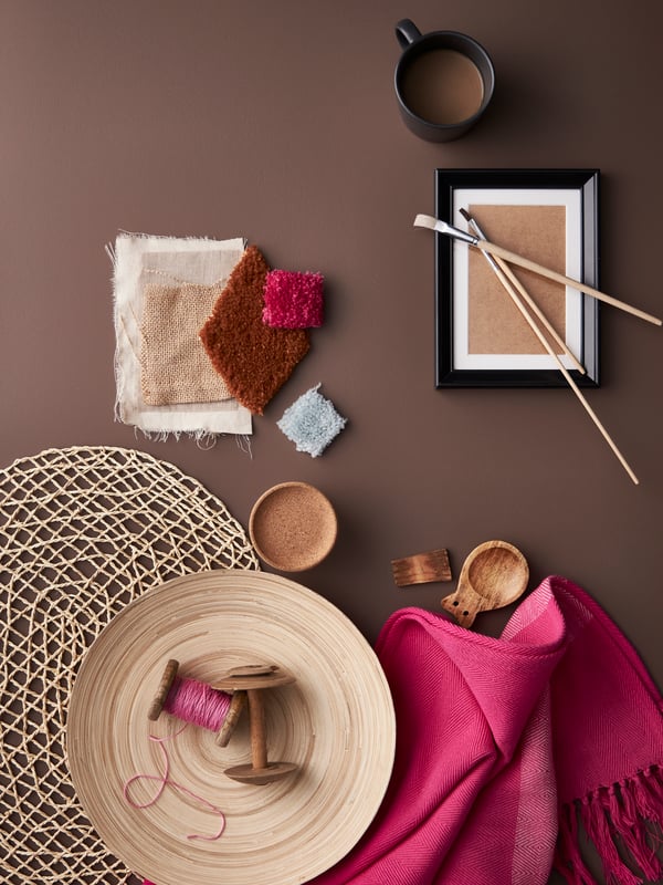 A HILLEGÄRD pink throw and a bamboo dish on a palm-leaf place mat, beside some fabric samples and a small black frame.