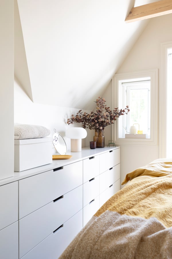 NORDLI kommode ved siden av en seng med en lampe, en vase med blomster og et fat på toppen.