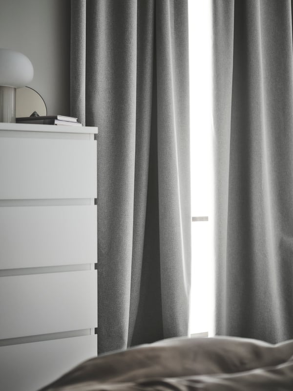A pair of grey HANNALENA room darkening curtains at a window by a white MALM chest of 6 drawers that has a DESJA lamp on it.